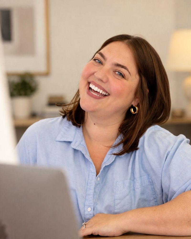Maxey laughing at the camera as she leans on a wooden table, an open laptop in the foreground. She wears a light blue button-up shirt and gold hoop earrings.